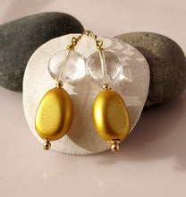 Load image into Gallery viewer, A pair of gold-tone dangle earrings with Clear Crystal Quartz beads and gold oval wooden drops. 14-karat Gold Filled findings they are displayed on a white background with two dark gray rocks behind it.