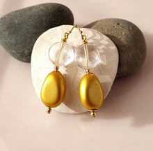 Load image into Gallery viewer, A pair of gold-tone dangle earrings with Clear Crystal Quartz beads and gold oval wooden drops. 14-karat Gold Filled findings they are displayed on a white background with two dark gray rocks behind it.