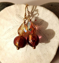 Load image into Gallery viewer, A pair of handmade dangle earrings with 14 karat gold filled ear wires, faceted amber-brown Swarovski Crystal beads, and round dark golden-brown Murano glass beads with metallic gold flecks, displayed on a textured white stone surface.