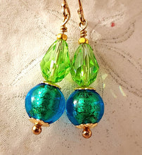 Load image into Gallery viewer, A pair of handmade drop earrings featuring faceted lime green Swarovski crystal beads, iridescent blue Murano glass beads, and 14 karat gold filled metal, displayed on a textured off-white fabric background.