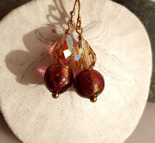 Load image into Gallery viewer, A pair of handmade dangle earrings with 14 karat gold filled ear wires, faceted amber-brown Swarovski Crystal beads, and round dark golden-brown Murano glass beads with metallic gold flecks, displayed on a textured white stone surface.