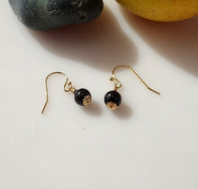 Load image into Gallery viewer, A pair of dangle earrings with large glossy black Onyx beads, textured oxidized 14 karat Gold-Filled findings and Gold fishhook wires, displayed on a white surface with a yellow flower in the background.