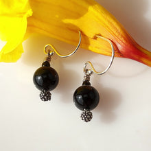 Load image into Gallery viewer, A pair of dangle earrings with large glossy black Onyx beads, textured oxidized sterling silver details, and Sterling Silver fishhook wires, displayed on a white surface with a yellow flower in the background.