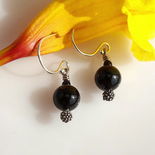 Load image into Gallery viewer, A pair of dangle earrings with large glossy black Onyx beads, textured oxidized sterling silver details, and Sterling Silver fishhook wires, displayed on a white surface with a yellow flower in the background.