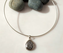 Load image into Gallery viewer, A sterling silver neck wire necklace with a teardrop-shaped Druzy pendant displayed on a white surface with smooth gray stones behind it.