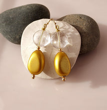 Load image into Gallery viewer, A pair of gold-tone dangle earrings with Clear Crystal Quartz beads and gold oval wooden drops. 14-karat Gold Filled findings they are displayed on a white background with two dark gray rocks behind it.
