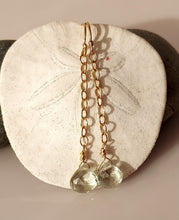 Load image into Gallery viewer, A pair of drop earrings with 14 karat gold-filled wire and findings with a dangling faceted green amethyst gemstone  displayed on a white sand dollar shell.
