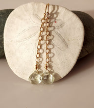Load image into Gallery viewer, A pair of drop earrings with 14 karat gold-filled wire and findings with a dangling faceted green amethyst gemstone  displayed on a white sand dollar shell.
