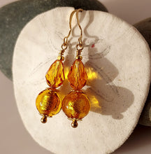 Load image into Gallery viewer, A pair of dangling earrings with faceted amber-gold Swarovski Crystal beads and textured gold Murano glass round beads, using 14 karat gold-filled findings hanging against a light textured background.

