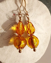 Load image into Gallery viewer, A pair of dangling earrings with faceted amber-gold Swarovski Crystal beads and textured gold Murano glass round beads, using 14 karat gold-filled findings hanging against a light textured background.
