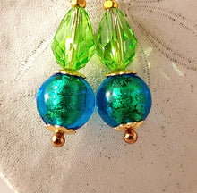 Load image into Gallery viewer, A pair of handmade drop earrings featuring faceted lime green Swarovski crystal beads, iridescent blue Murano glass beads, and 14 karat gold filled metal, displayed on a textured off-white fabric background.
