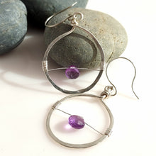Load image into Gallery viewer, A pair of handmade silver hoop earrings with thin wire details and small purple amethyst gemstones, displayed on a white surface next to a gray stone.
