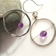 Load image into Gallery viewer, A pair of handmade silver hoop earrings with thin wire details and small purple amethyst gemstones, displayed on a white surface next to a gray stone.
