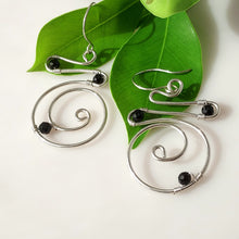 Load image into Gallery viewer, A pair of handmade sterling silver spiral wire hoop earrings with hand wire-wrapped black onyx beads, displayed on vibrant green leaves against a clean white background.
