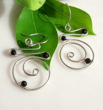 Load image into Gallery viewer, A pair of handmade sterling silver spiral wire hoop earrings with hand wire-wrapped black onyx beads, displayed on vibrant green leaves against a clean white background.
