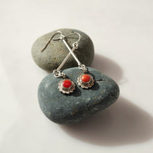 Load image into Gallery viewer, A pair of Sterling Silver dangle earrings with round Carnelian stones in a scalloped floral setting, displayed on smooth dark gray stones against a light neutral background.
