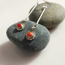 Load image into Gallery viewer, A pair of Sterling Silver dangle earrings with round Carnelian stones in a scalloped floral setting, displayed on smooth dark gray stones against a light neutral background.
