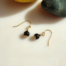 Load image into Gallery viewer, A pair of dangle earrings with large glossy black Onyx beads, textured oxidized 14 karat Gold-Filled findings and Gold fishhook wires, displayed on a white surface with a yellow flower in the background.
