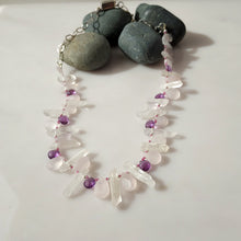 Load image into Gallery viewer, A beaded necklace made of clear quartz crystal points and pale pink rose quartz beads, accented with small purple amethyst beads, displayed on a white surface with dark gray decorative stones.

