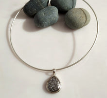 Load image into Gallery viewer, A sterling silver neck wire necklace with a teardrop-shaped Druzy pendant displayed on a white surface with smooth gray stones behind it.
